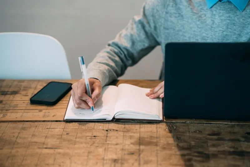 Hands writing questions in a notebook beside a laptop and a phone on a wooden table